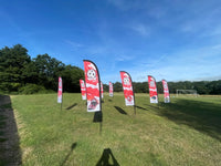 A row of red and white feather flag banners with football themed graphics, displayed on a grassy field under a blue sky with trees in the background.